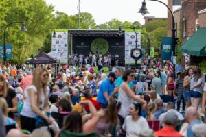 A crowd of people are sitting and standing in a downtown street that leads to a stage with a musical group performing in front of Friday Night on White sign.